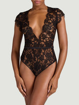 A front view of a model wearing the Aphrodite bodysuit. Made from a black, sheer French Leavers lace with a silk detail around the waistband and a silk covered button at the back. It features a plunge neckline and delicate capped sleeves both showcasing the beautiful eyelash edge of the lace.