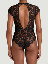 A back view of a model wearing the Aphrodite bodysuit. Made from a black, sheer French Leavers lace with a silk detail around the waistband and a silk covered button at the back. It features a delicate capped sleeve showcasing the beautiful eyelash edge of the lace and a back cut out highlighting the elegance of the spine.