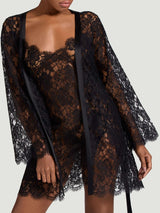 The front view of a model wearing the Coco de Mer Aphrodite slip and Aphrodite robe. Made from a black, sheer French Leavers lace with a delicate eyelash edge along the neck and hemlines and along the cuffs and hemline on the robe. Featuring a side split and delicate thin straps on the slip and a luxurious black silk front edge and tie on the robe.