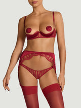 A model wearing an intricate red French Leaver's lace and luxurious silk suspender knicker with a high waist and adjustable straps, featuring golden hardware and scallop edge lace details. This is paired with a red quarter cup bra featuring details of scallop edged Leaver's Lace lining a silk covered quarter cup and silk straps with a hook and eye closure at the spine.