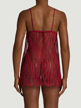 A back view of a model wearing a short red slip made from a delicate red French leaver's lace with a hemline featuring a scalloped edge lace detail. There is a red silk under band, shoulder straps and a red silk covered button.