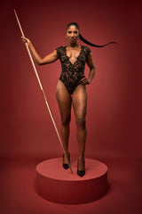 A front view of a model wearing the Aphrodite bodysuit. Made from a black, sheer French Leavers lace with a silk detail around the waistband and a silk covered button at the back. It features a plunge neckline and delicate capped sleeves both showcasing the beautiful eyelash edge of the lace.