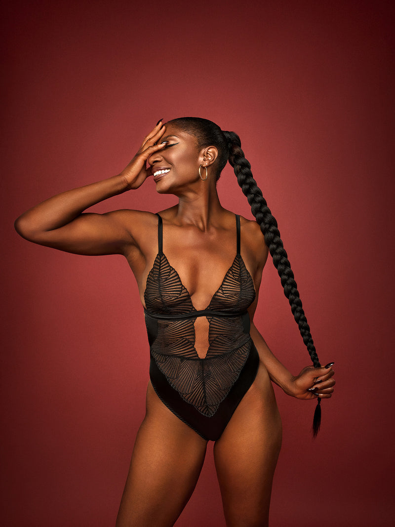 Dame Denise Lewis wears a black Coco de Mer Athena Bodysuit with graphic embroidery and a deep plunging neckline. It includes fan edged cups, an underbust band of jet black silk, adjustable silk straps, and gold hook and loop fastenings