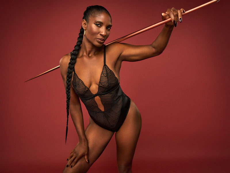 Dame Denise Lewis wears a black Coco de Mer Athena Bodysuit with graphic embroidery and a deep plunging neckline. It includes fan edged cups, an underbust band of jet black silk, adjustable silk straps, and gold hook and loop fastenings