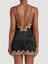 Model showing the back of a Coco de Mer 'Reina' black silk slip. The garment has a gold embroidered lace trim, silk-covered straps and a deep V low back. 