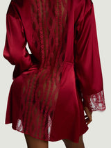 A close up view of a model wearing the Coco de Mer Renata robe. This is a red silk robe featuring a panel of red French leaver's lace down the centre back and along the bottom cuffs of the sleeve. There is a red silk tie that creates a flattering shape at the waist when tied.